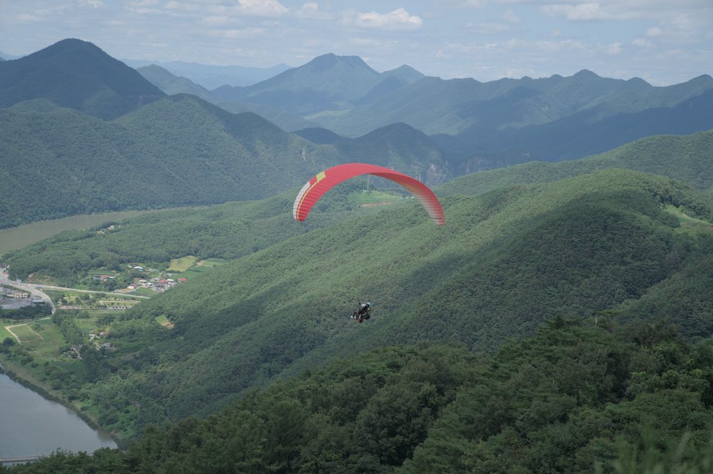 Parapente à voile rouge survolant des montagnes boisées et une vallée fluviale — raw file