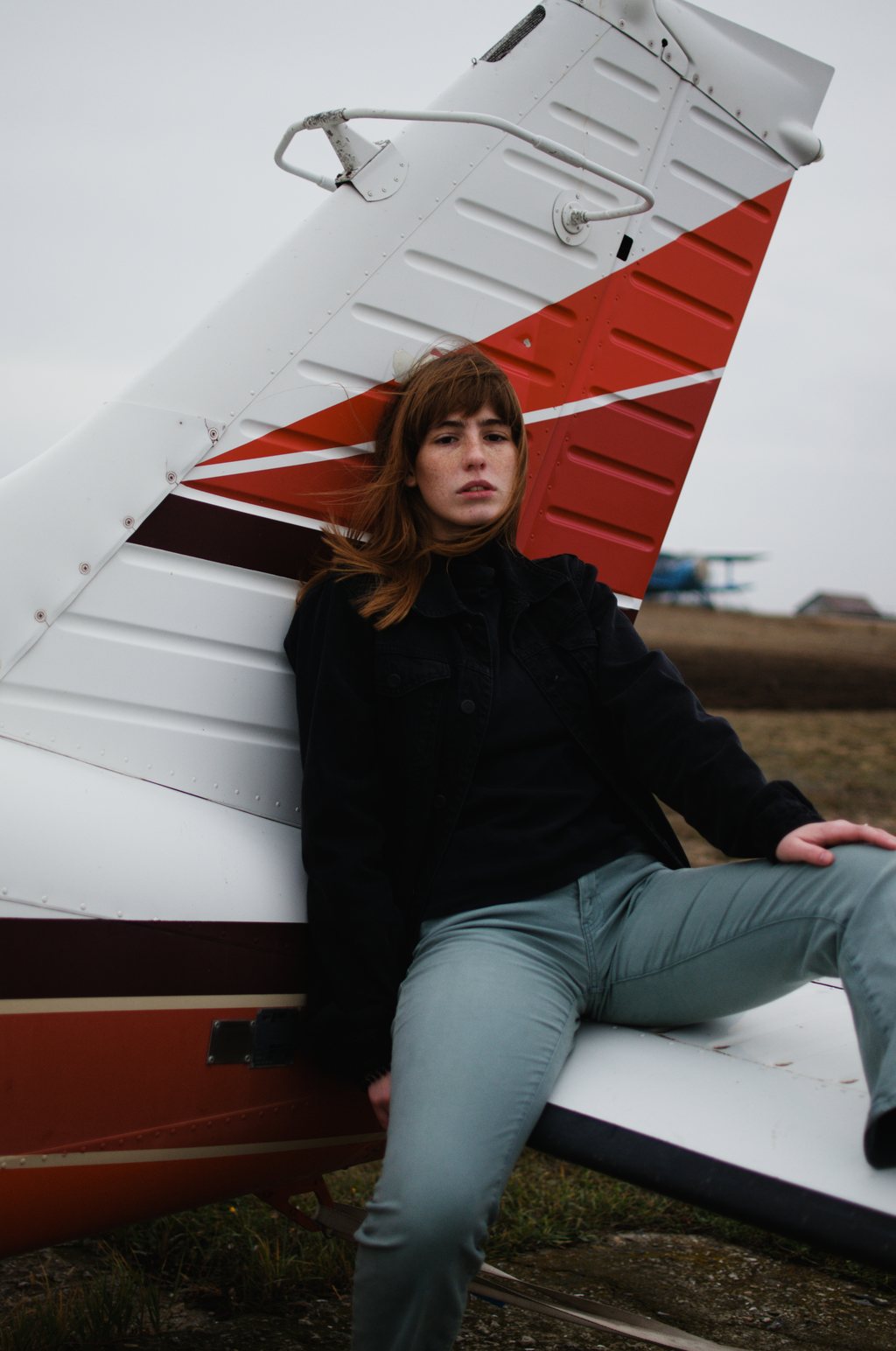 Portrait, jeune femme aux cheveux auburn en veste noire, adossée à l'empennage rouge et blanc d'un petit avion sur un aérodrome au ciel couvert — neochrome version