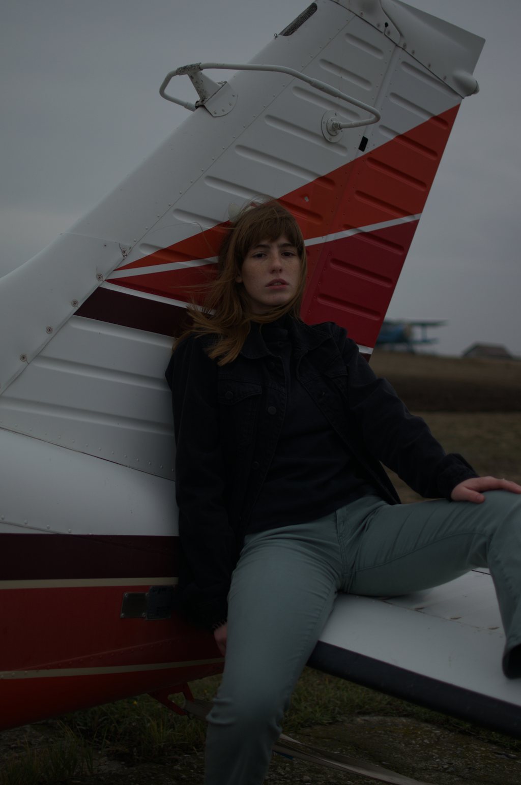 Portrait, jeune femme aux cheveux auburn en veste noire, adossée à l'empennage rouge et blanc d'un petit avion sur un aérodrome au ciel couvert — raw file