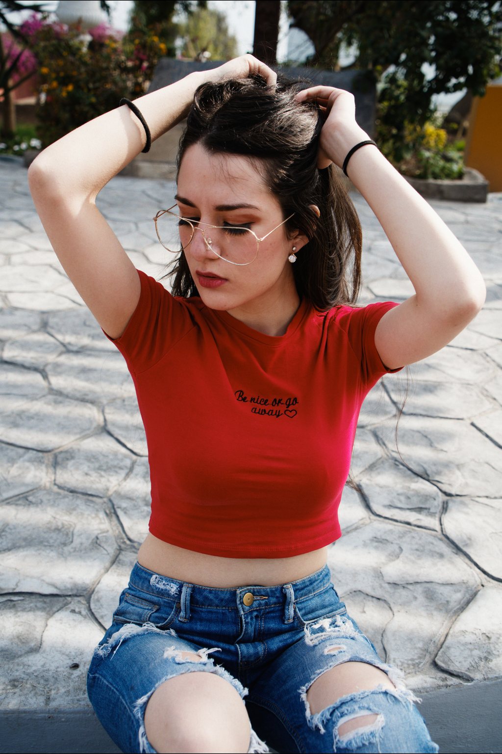 Portrait, jeune femme à lunettes en t-shirt rouge et jean déchiré, assise sur un sol pavé, mains dans les cheveux — neochrome version