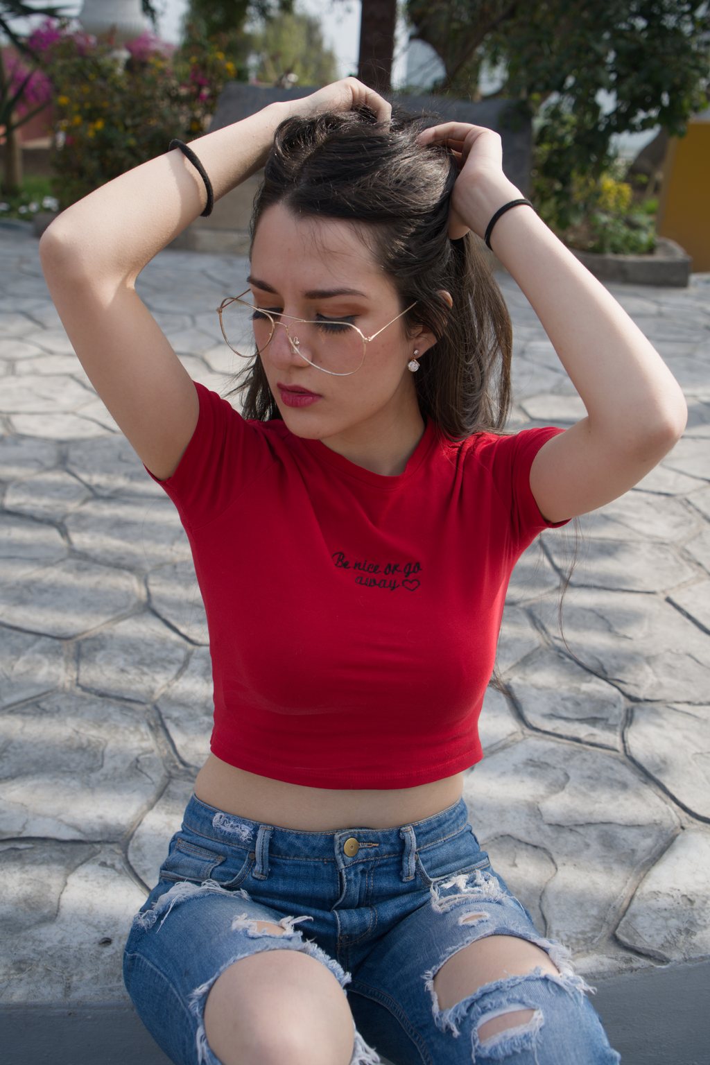 Portrait, jeune femme à lunettes en t-shirt rouge et jean déchiré, assise sur un sol pavé, mains dans les cheveux — raw file