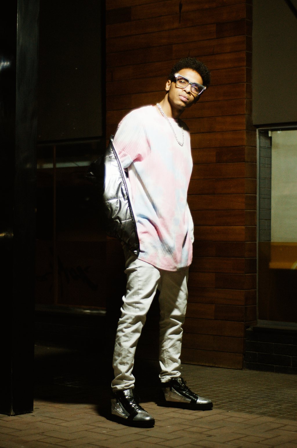 Portrait de nuit, jeune homme à lunettes adossé à un mur de briques, pull tie-dye pastel et doudoune argentée sur l'épaule — neochrome version