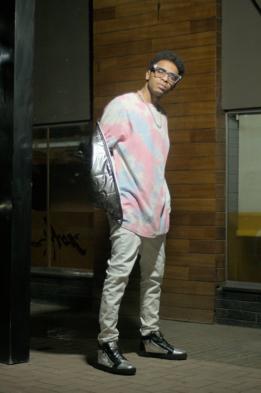 Portrait de nuit, jeune homme à lunettes adossé à un mur de briques, pull tie-dye pastel et doudoune argentée sur l'épaule — raw file