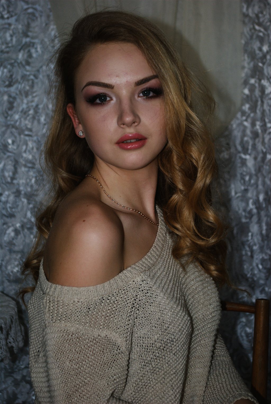 Portrait, jeune femme aux longs cheveux blonds ondulés, pull beige tombant sur l'épaule, devant une tenture grise à motifs — neochrome version