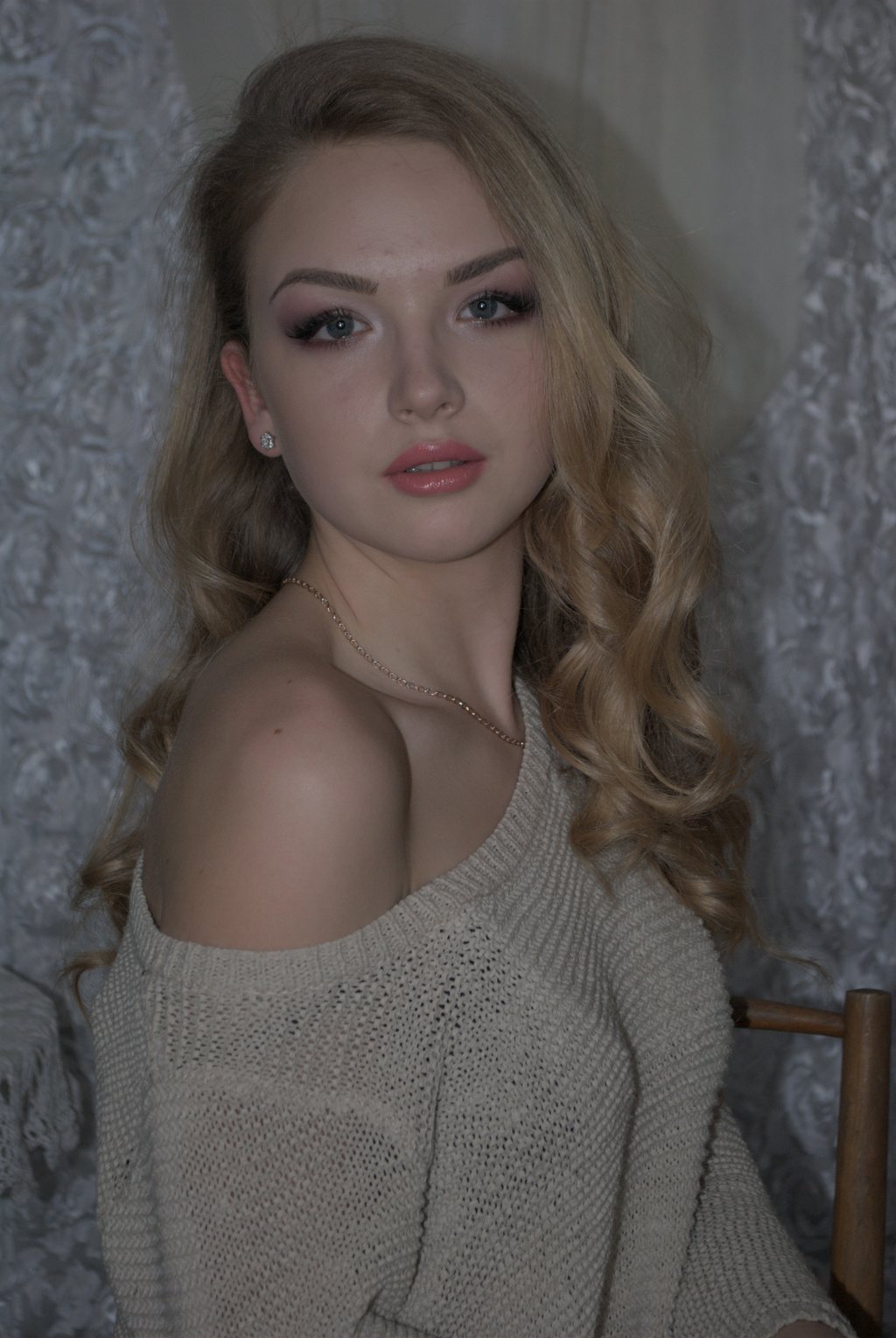 Portrait, jeune femme aux longs cheveux blonds ondulés, pull beige tombant sur l'épaule, devant une tenture grise à motifs — raw file