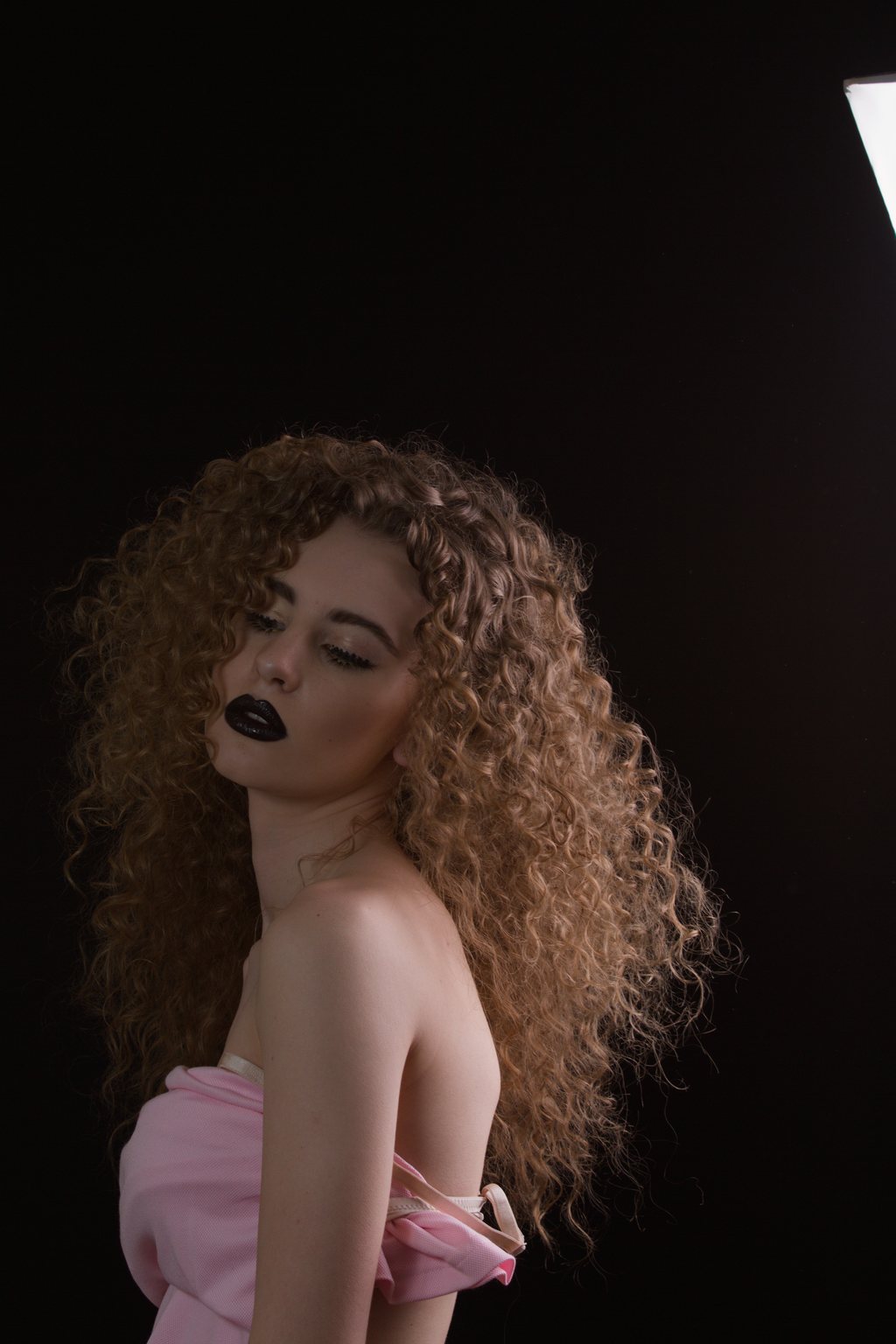 Portrait studio, jeune femme aux cheveux bouclés volumineux, rouge à lèvres noir, bustier rose sur fond noir — raw file