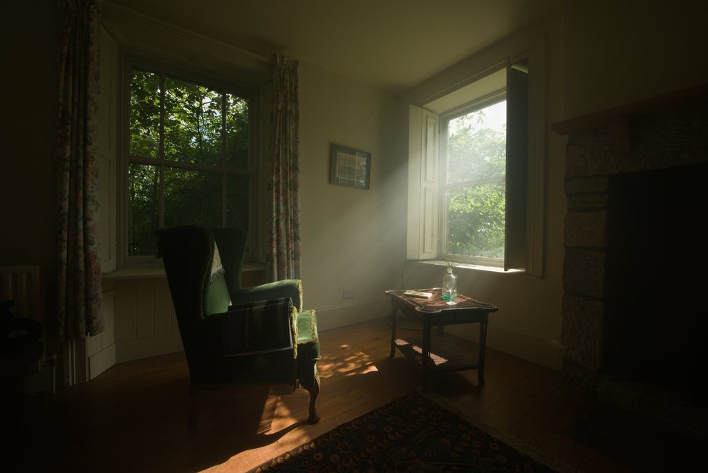 Intérieur de maison tamisé, fauteuil à oreilles en velours vert et guéridon de bois près d'une fenêtre à petits carreaux, rideaux fleuris, rai de lumière traversant la pièce jusqu'au parquet — raw file