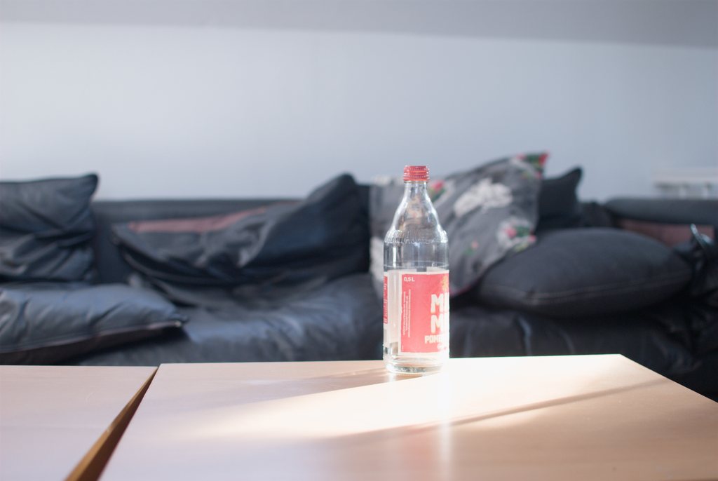 Bouteille d'eau minérale en verre à l'étiquette rouge, posée dans un rai de lumière sur une table en bois, canapé de cuir sombre et coussins flous à l'arrière-plan — raw file